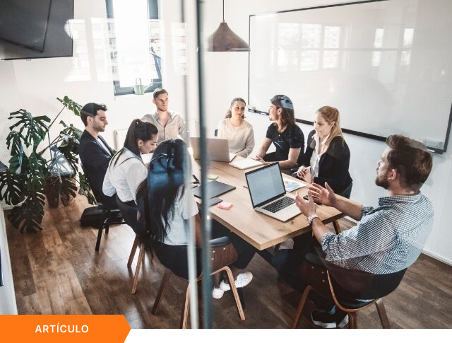 Imagen de portada para artículo que muestra una vista desde arriba de un equipo diverso en una reunión estratégica alrededor de una mesa de madera, vista a través de una pared de cristal, con una etiqueta naranja de ARTÍCULO en la esquina inferior izquierda.
