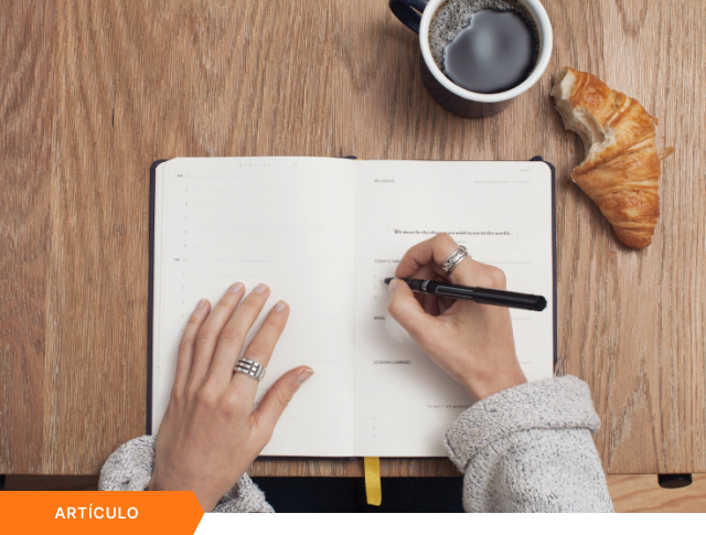 Portada de artículo que muestra una vista cenital de una persona escribiendo en una agenda sobre una mesa de madera con café y un croissant, con etiqueta naranja de ARTÍCULO en la esquina inferior.