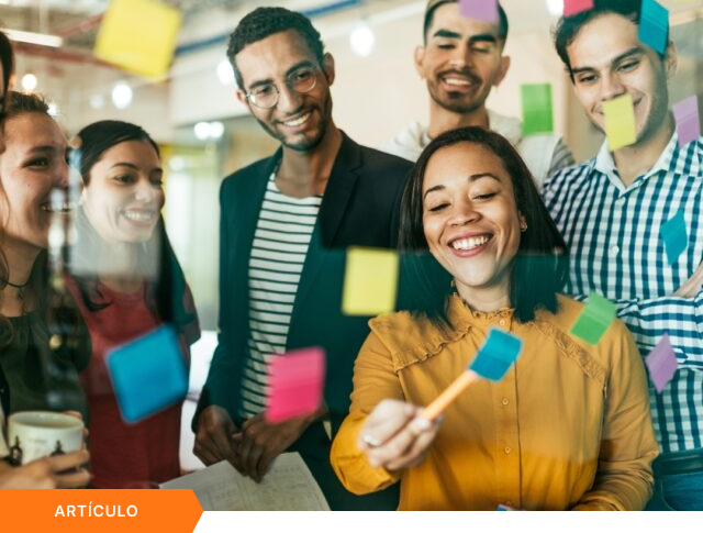 Imagen de portada para artículo mostrando a un grupo diverso de siete profesionales sonriendo y pegando notas adhesivas en un panel de cristal, con una etiqueta naranja de ARTÍCULO en la esquina inferior izquierda.