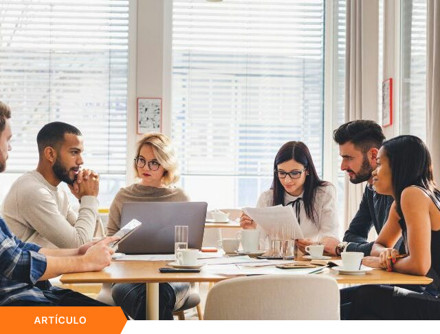 Imagen de portada para artículo mostrando a un equipo de seis profesionales en una reunión estratégica alrededor de una mesa de madera, con una etiqueta naranja que dice ARTÍCULO en la esquina inferior izquierda.