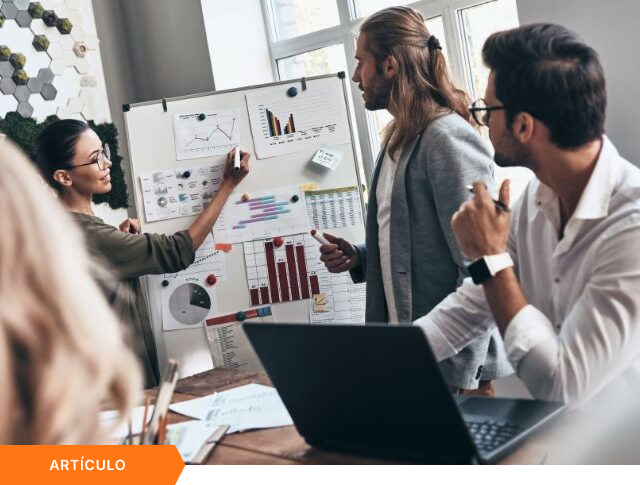 Portada de artículo que muestra a una profesional explicando gráficos y métricas en una pizarra durante una reunión, con etiqueta naranja de ARTÍCULO en la esquina inferior.