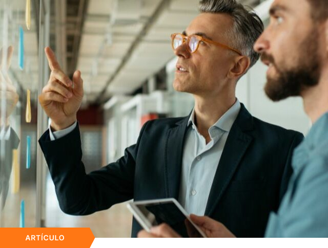 Imagen de portada para artículo que muestra a un ejecutivo senior con gafas naranja explicando una estrategia en un panel de notas adhesivas a un colega con tablet, con una etiqueta naranja de ARTÍCULO en la esquina inferior izquierda.