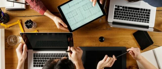 Vista superior de un equipo multidisciplinario trabajando en diseño técnico con laptops y tablets, con etiqueta naranja de ARTÍCULO en la esquina inferior.