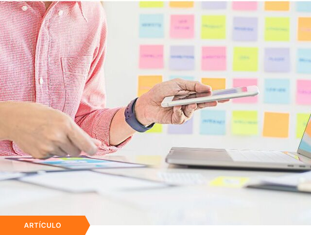 Imagen de portada para artículo mostrando a un profesional utilizando un smartphone y una laptop simultáneamente frente a una pizarra con notas adhesivas, con una etiqueta naranja de ARTÍCULO en la esquina inferior izquierda.