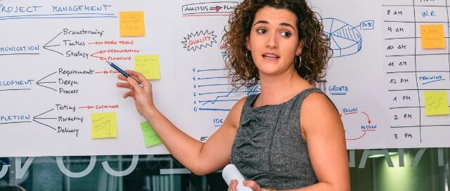 Imagen de portada para artículo mostrando a una profesional explicando un esquema de Project Management en una pizarra, con una etiqueta naranja de ARTÍCULO en la esquina inferior izquierda.