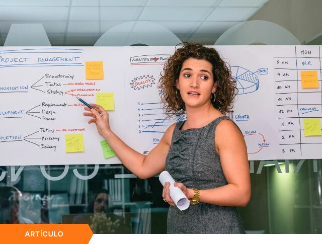 Imagen de portada para artículo mostrando a una profesional explicando un esquema de Project Management en una pizarra, con una etiqueta naranja de ARTÍCULO en la esquina inferior izquierda.