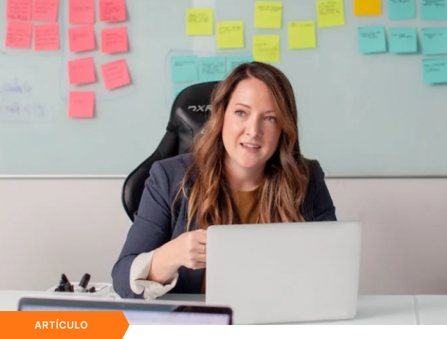 Portada de artículo que muestra a una profesional liderando una reunión con laptop frente a una pizarra con notas adhesivas de colores, con etiqueta naranja de ARTÍCULO en la esquina inferior.
