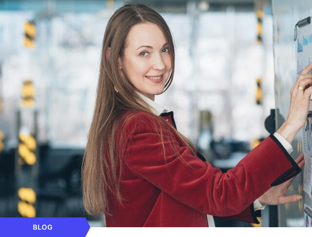 Mujer profesional sonriente con chaqueta roja apuntando a gráficos de rendimiento en un tablero de cristal, diseñada como imagen de portada para un artículo de blog sobre liderazgo, análisis de datos y seguimiento de métricas en la gestión de proyectos.