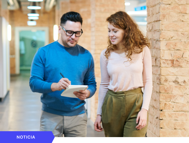 Portada de noticia que muestra a un hombre con gafas anotando en una libreta mientras camina y conversa con una colega en un pasillo de oficina, con etiqueta azul de NOTICIA en la esquina inferior.
